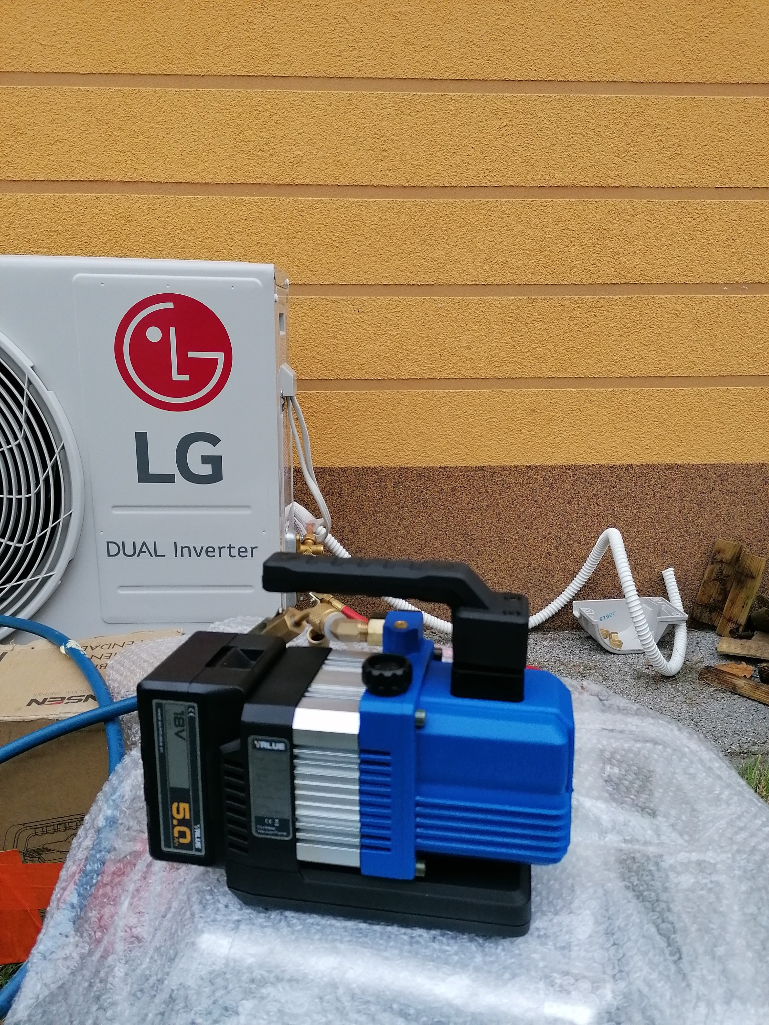 Pompa próżniowa VALUE 18V 5.0, podłączona do zewnętrznej jednostki klimatyzacji LG Dual Inverter, stojąca na folii bąbelkowej na tle żółtej ściany budynku.