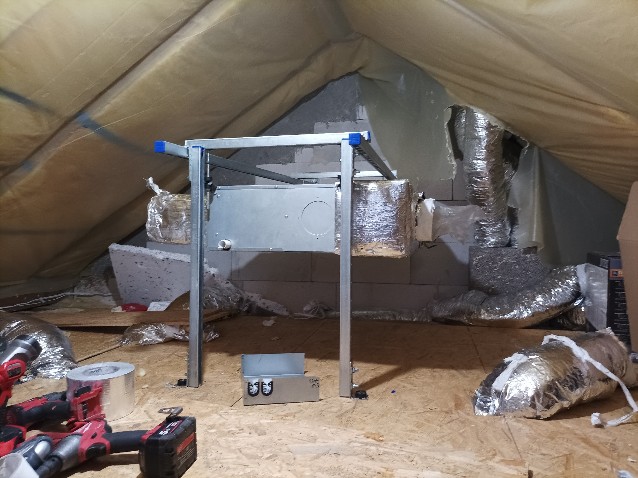 Instalacja centrali wentylacyjnej z rekuperacją w przestrzeni poddasza, widoczne kanały wentylacyjne w izolacji aluminiowej, narzędzia montażowe i elementy konstrukcyjne.