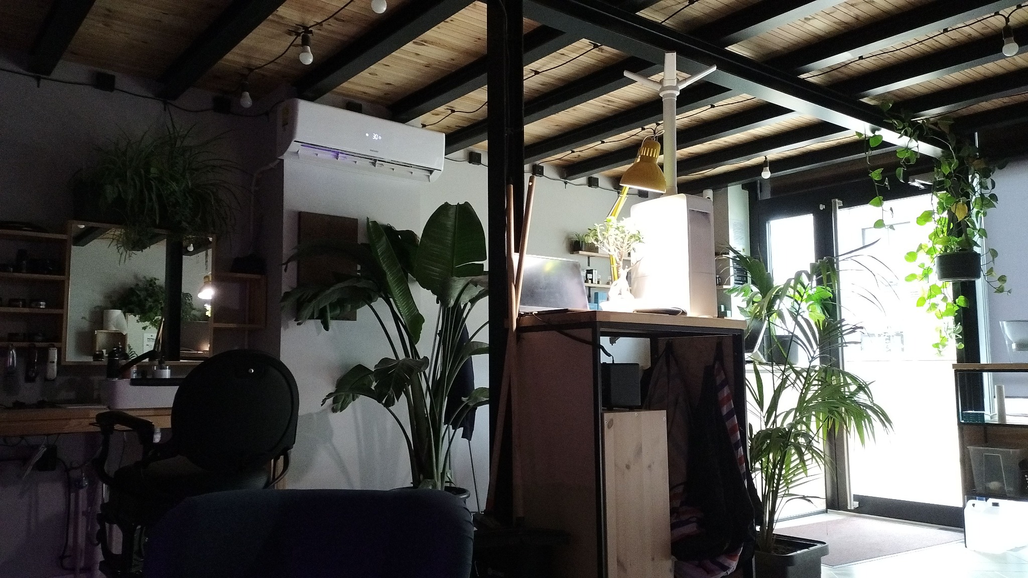 Wnętrze biura z zainstalowanym klimatyzatorem ściennym, widoczne rośliny doniczkowe, biurko z lampką i krzesło fryzjerskie w tle.