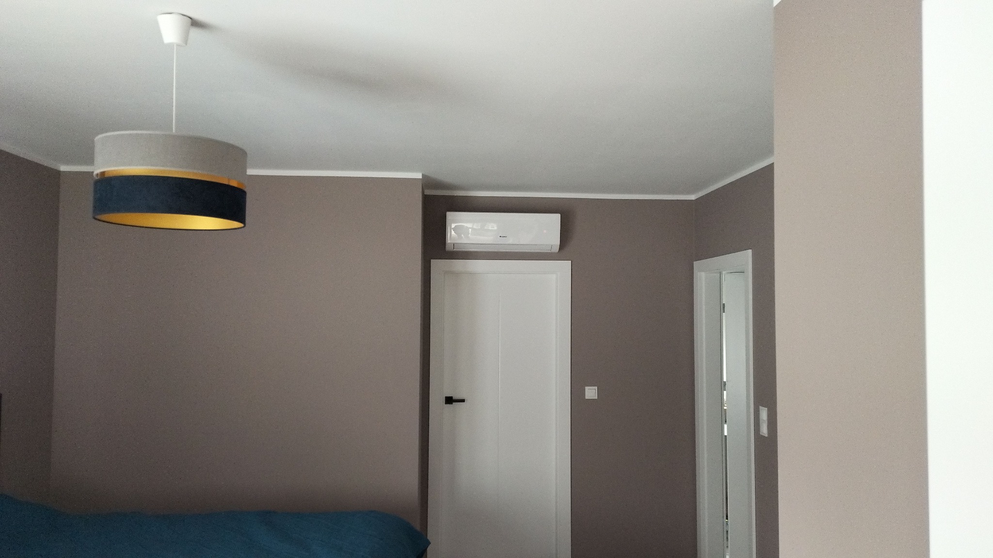 Wnętrze pokoju z zamontowaną białą jednostką klimatyzacji ściennej nad białą, zamkniętą drzwiami, z widoczną lampą sufitową z szarym i granatowym abażurem.