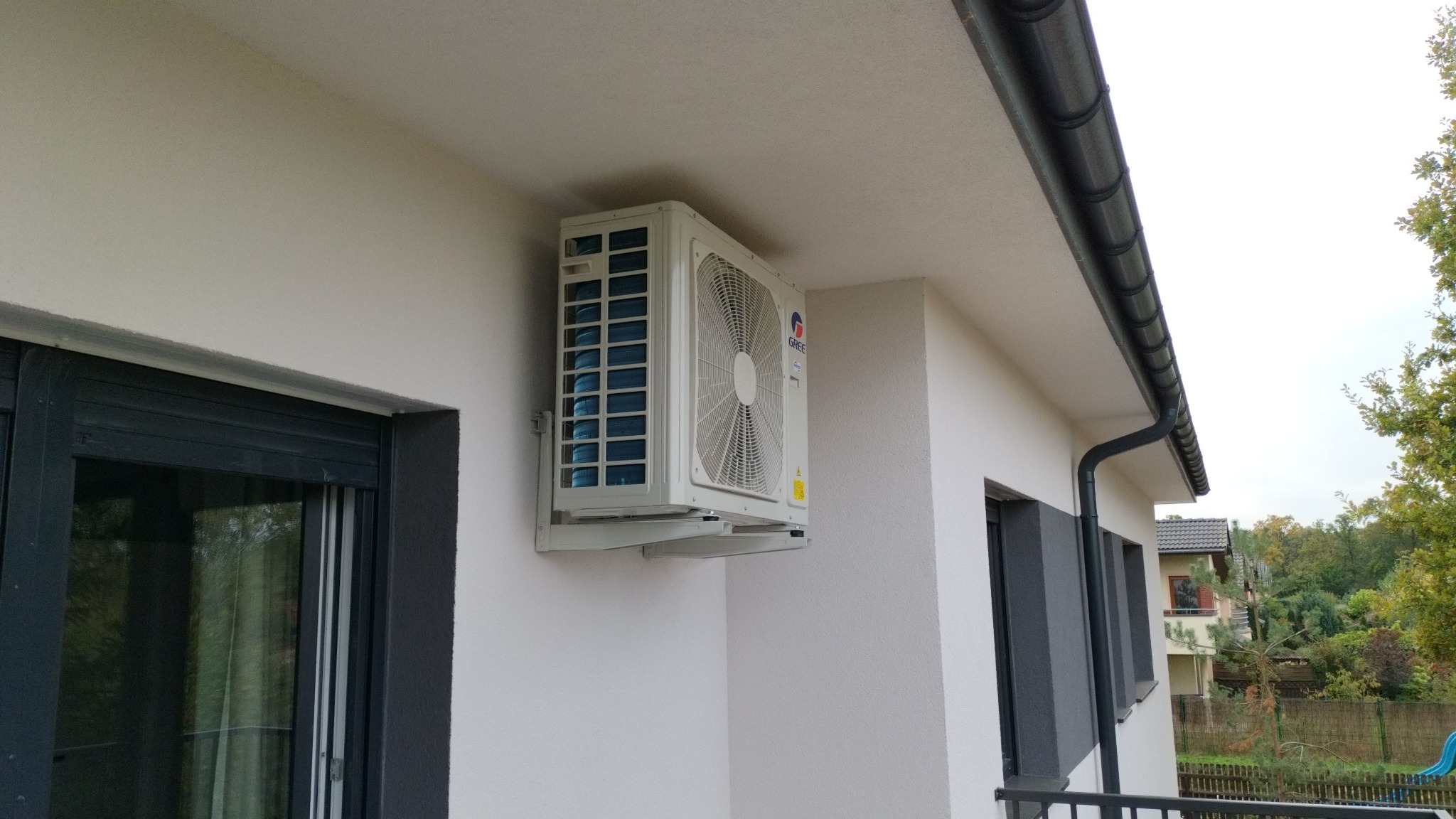 Zewnętrzna jednostka klimatyzacji marki Gree zamontowana na białej ścianie budynku, widoczna z boku, z fragmentem okna z ciemną ramą i rynną w tle.