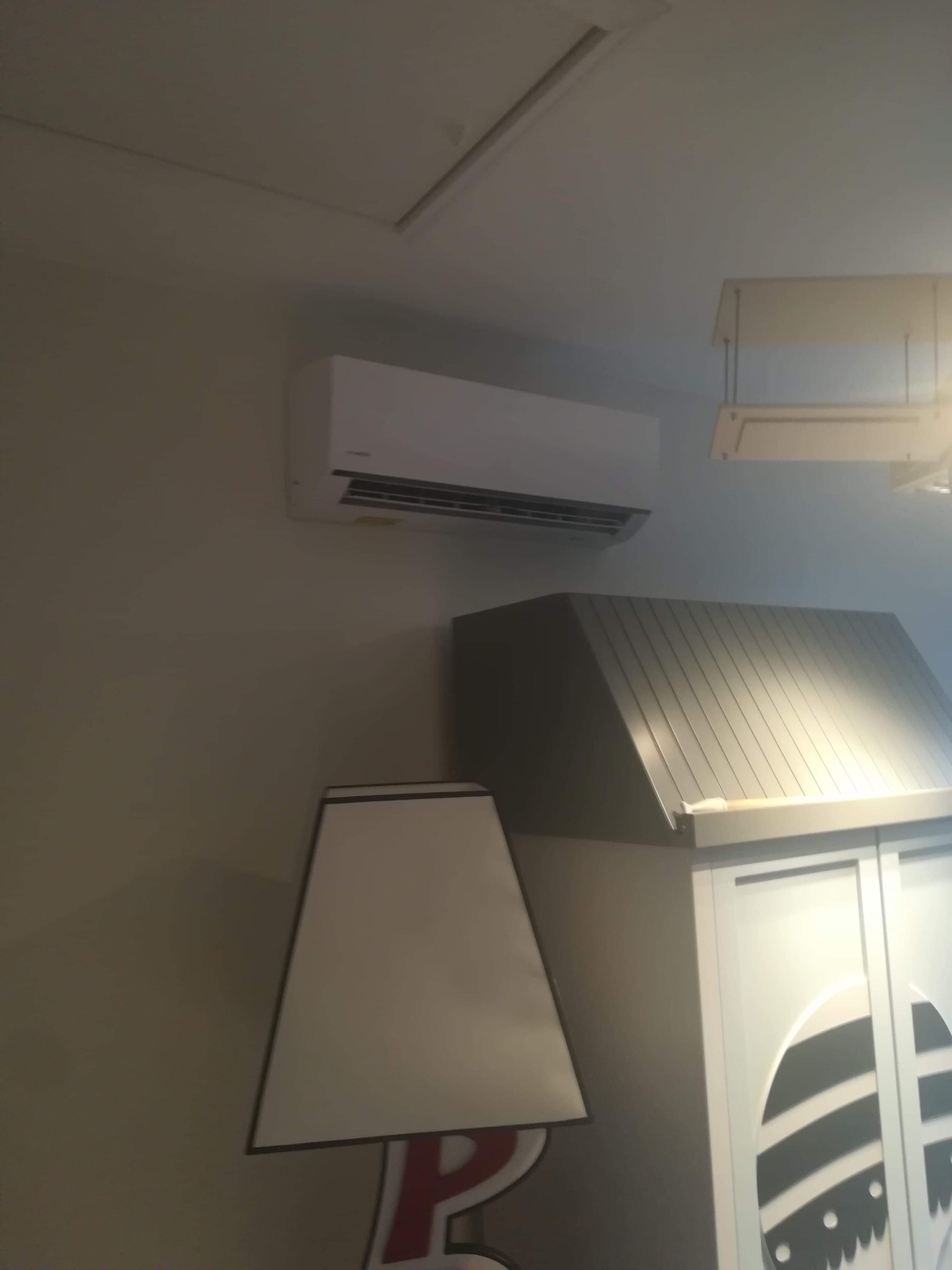 Biała jednostka klimatyzacyjna zamontowana na ścianie w pomieszczeniu, widoczna nad szafką z dekoracyjnym wzorem jajka i lampą stołową z czerwonym akcentem.