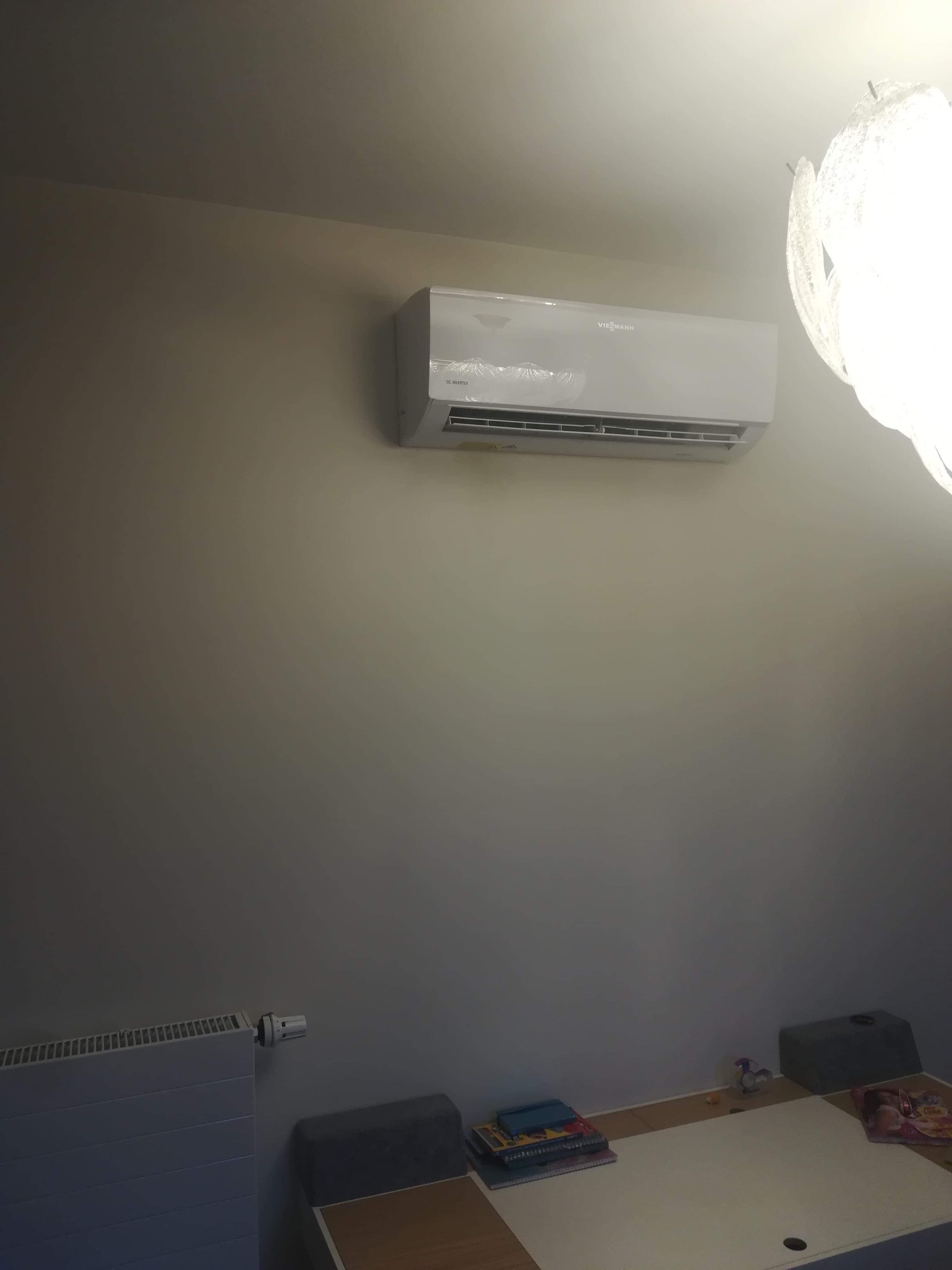 Biała klimatyzacja Viessmann zamontowana na szarej ścianie w pokoju, widoczny fragment dekoracyjnego żyrandola i biurka z książkami.
