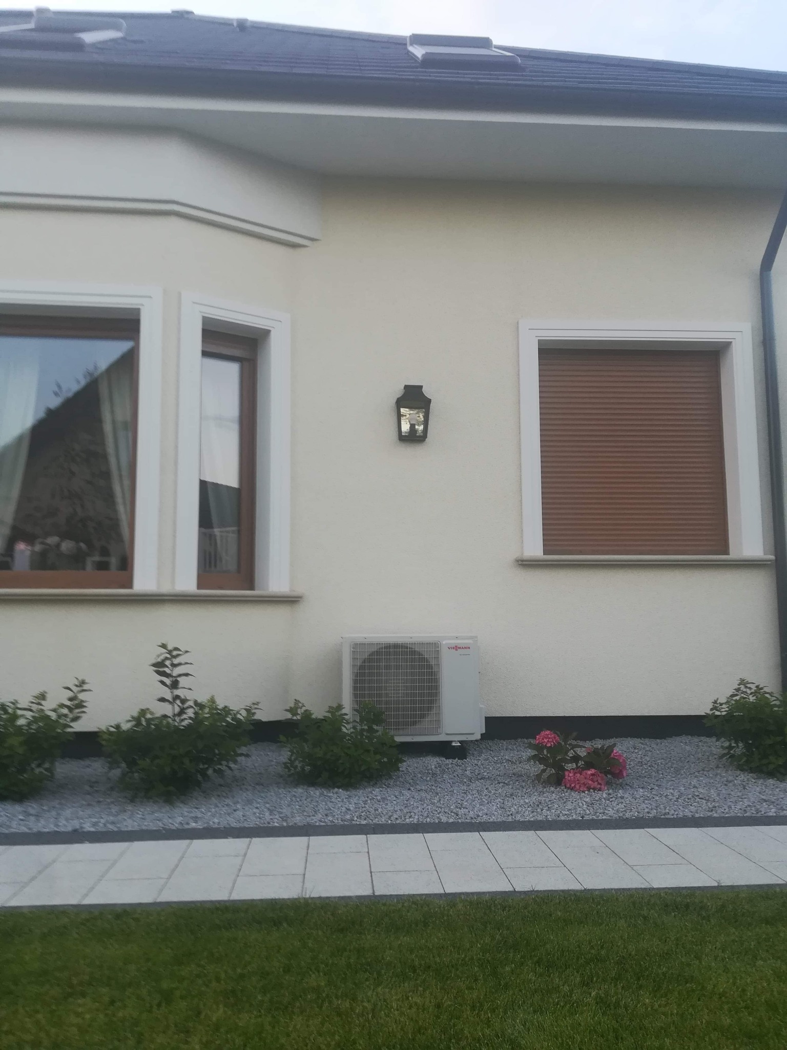 Biała jednostka zewnętrzna pompy ciepła Viessmann zamontowana na elewacji domu z jasną fasadą, widoczna lampa ścienna, okna z brązowymi roletami i dekoracyjny klomb z szarym żwirem i różowymi...