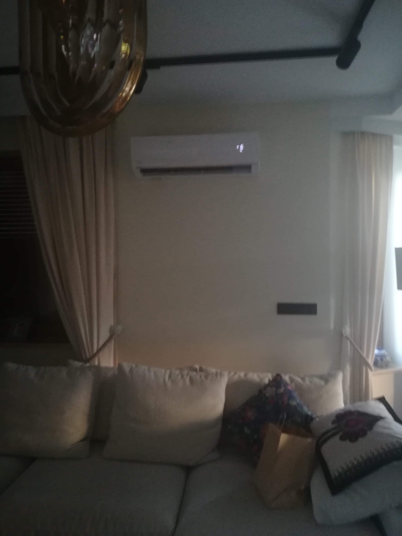 Biała jednostka wewnętrzna klimatyzacji zamontowana na ścianie nad kanapą w salonie, widoczna z perspektywy żyrandola.