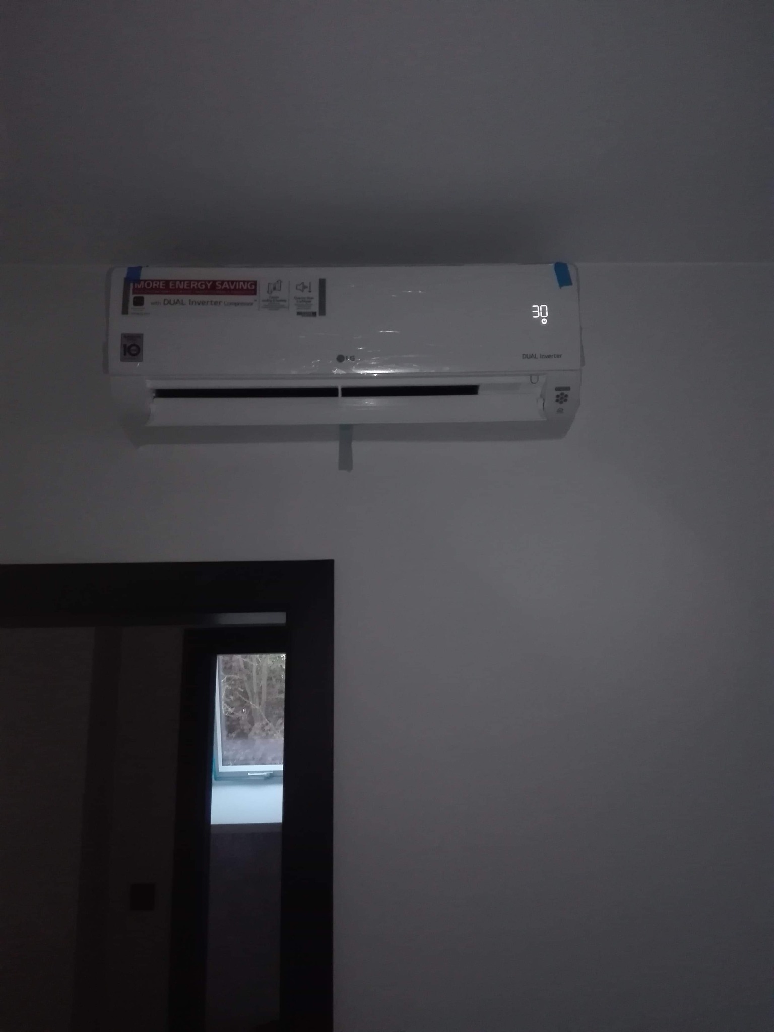 Biała jednostka klimatyzacji LG Dual Inverter zamontowana na ścianie w pokoju, nadal zabezpieczona folią ochronną, wyświetla temperaturę 30 stopni Celsjusza.