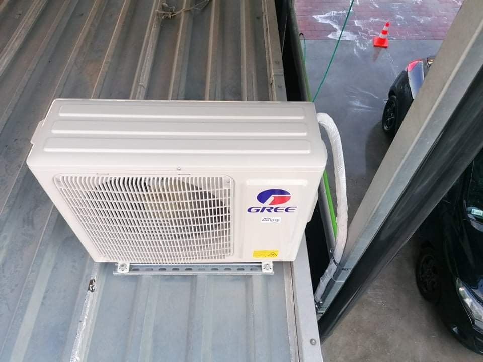 Biała jednostka zewnętrzna klimatyzacji marki Gree zamontowana na metalowej konstrukcji przy ścianie budynku, widoczne kable i rury instalacyjne, w tle fragment samochodu i pachołek drogowy.