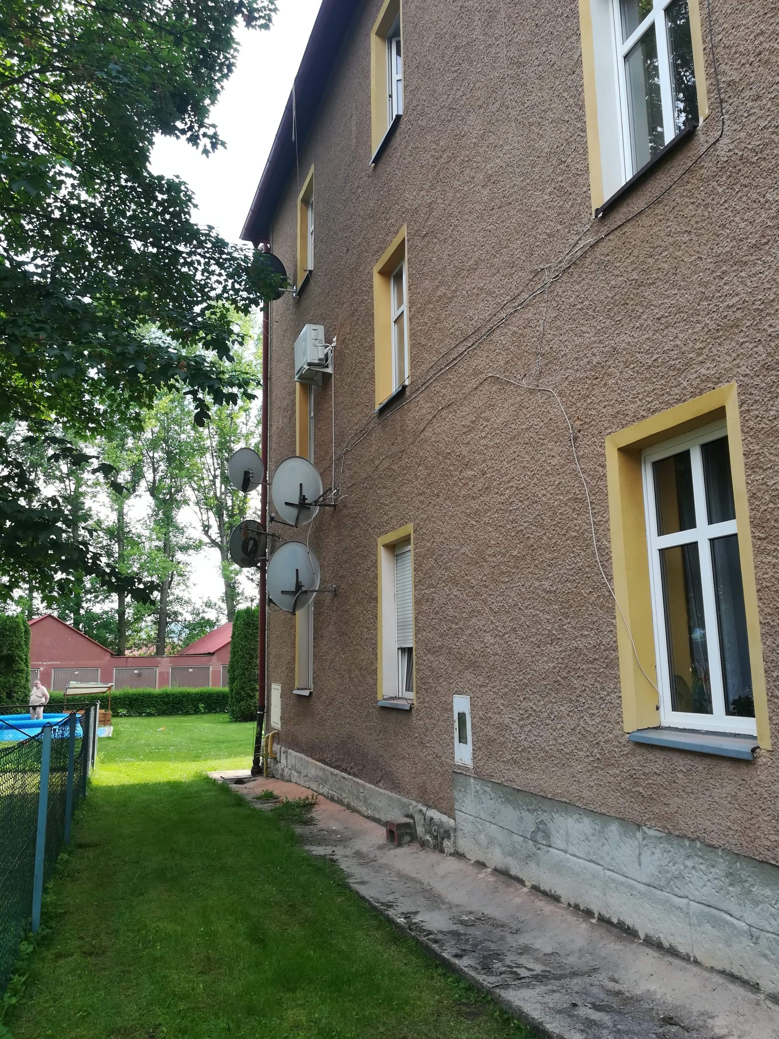 Elewacja budynku z zamontowanym klimatyzatorem i kilkoma antenami satelitarnymi, widok z boku na tle zielonego trawnika i drzew.