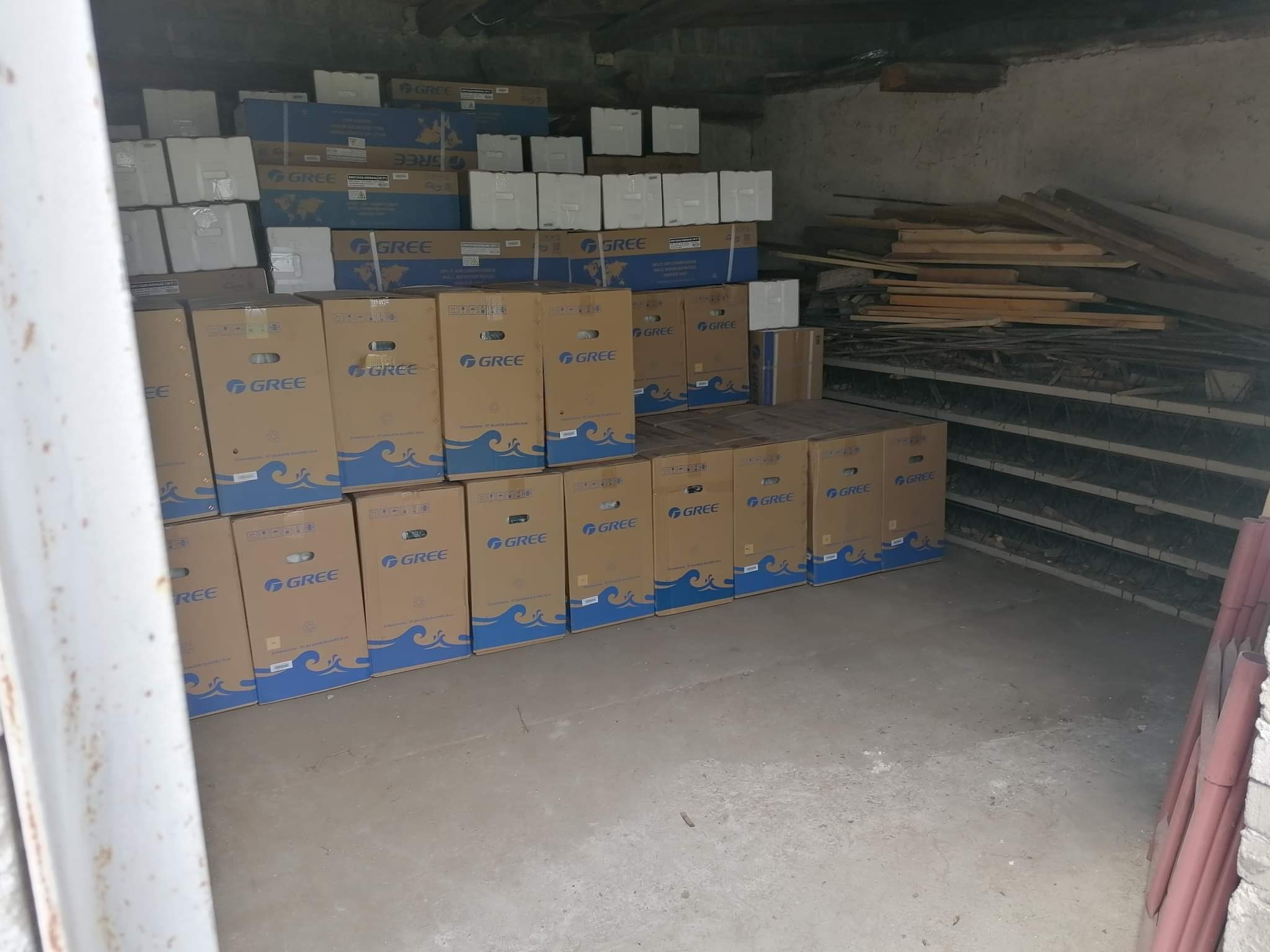 Stos kartonowych pudeł z logo GREE, prawdopodobnie klimatyzatorów lub pomp ciepła, składowanych w piwnicy lub magazynie.