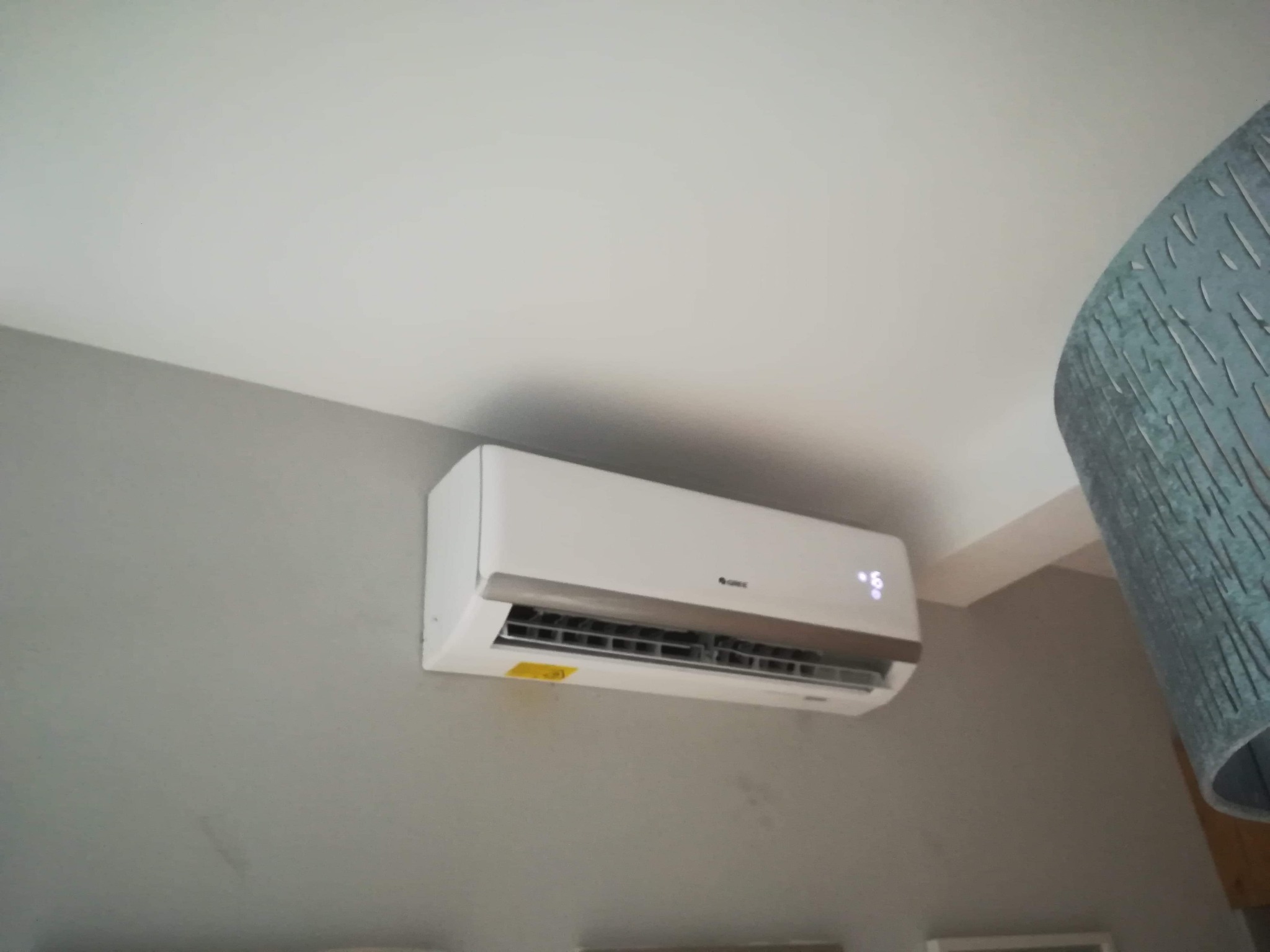 Biały klimatyzator ścienny marki Gree zamontowany w pomieszczeniu z szarą ścianą i białym sufitem, widoczny fragment lampy z boku kadru.