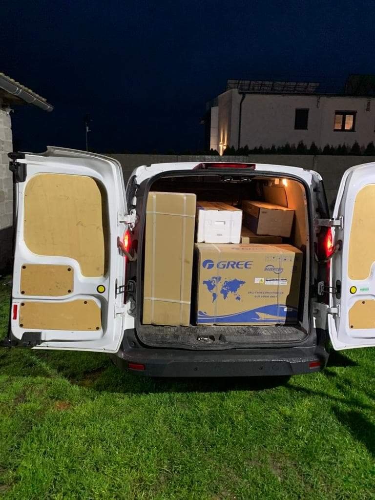 Tył białego samochodu dostawczego z otwartymi drzwiami, wypełnionego kartonami, w tym pudełkiem z logo GREE i napisem 'INVERTER', na tle trawnika i domu w nocy.