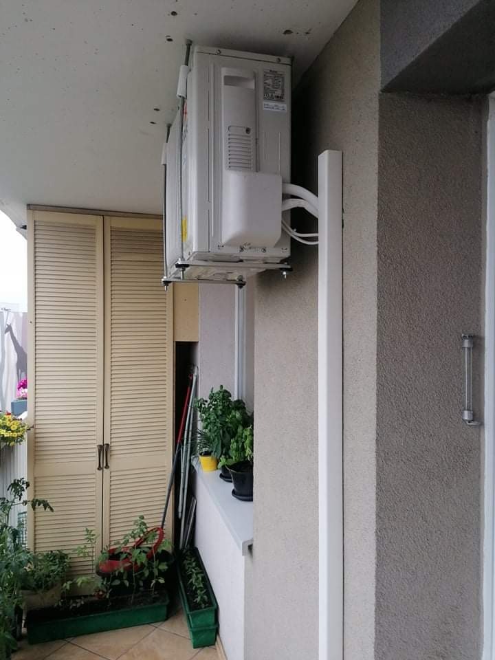Zewnętrzna jednostka klimatyzatora zamontowana pod sufitem balkonu, z widocznymi przewodami i elementami montażowymi, na tle ściany z roślinami w doniczkach i szafką balkonową.