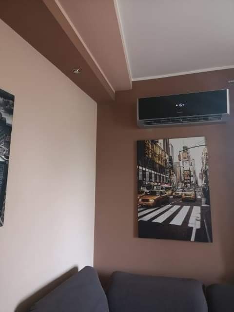 Czarny klimatyzator ścienny w nowoczesnym salonie z obrazem nowojorskiej ulicy, widoczny fragment szarej sofy.