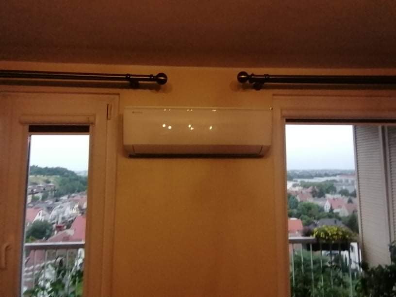 Biała jednostka klimatyzacji zamontowana na ścianie nad oknem balkonowym z widokiem na miasto w tle. Widoczne karnisze nad oknami.