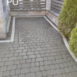 Ułożona szara kostka brukowa z jasną obwódką, widok na bramę z poziomymi przęsłami i tujami po prawej stronie. Zdjęcie wykonane po realizacji.