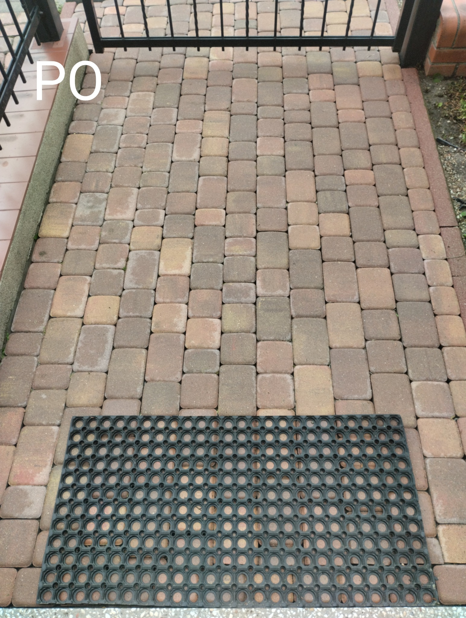 Ułożona kostka brukowa w odcieniach brązu i beżu, z czarną wycieraczką u dołu kadru, widok z góry, fragment metalowej barierki w tle.