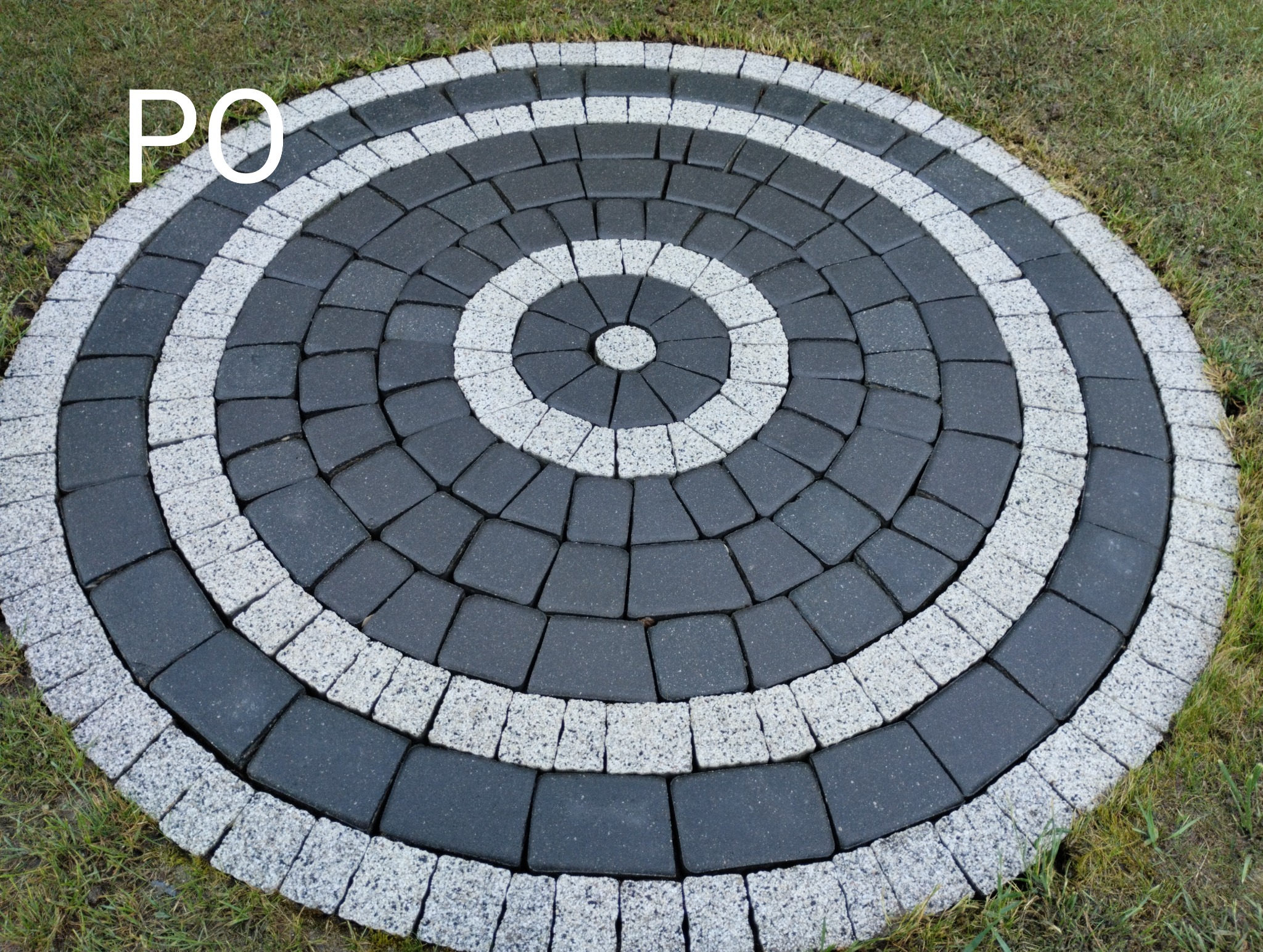 Okrągła kompozycja z kostki brukowej w odcieniach szarości i bieli, otoczona trawnikiem.