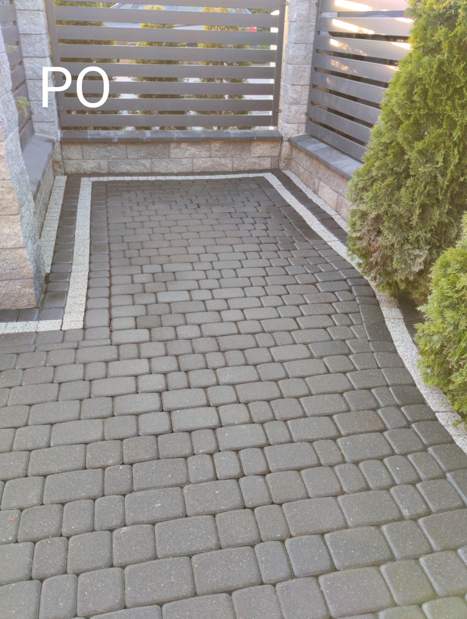 Ułożona szara kostka brukowa z jasną obwódką, widok na bramę z poziomymi przęsłami i tujami po prawej stronie. Zdjęcie wykonane po realizacji.