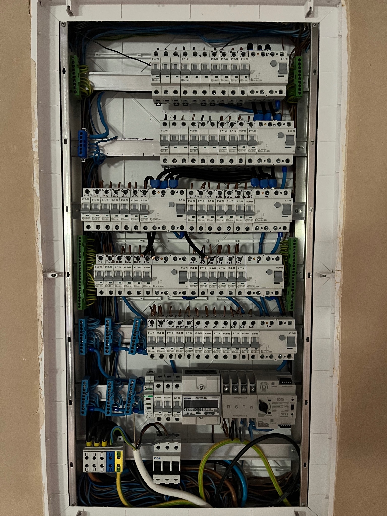 Rozdzielnica elektryczna w białej obudowie, widoczne liczne bezpieczniki, kable w różnych kolorach i złączki. Instalacja wykonana schludnie i profesjonalnie.