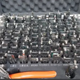 Otwarty futerał z zestawem metalowych kluczy/próbek do zamków samochodowych, ułożonych w rzędach, z pomarańczowym narzędziem widocznym na dole, na tle ceglanej ściany.