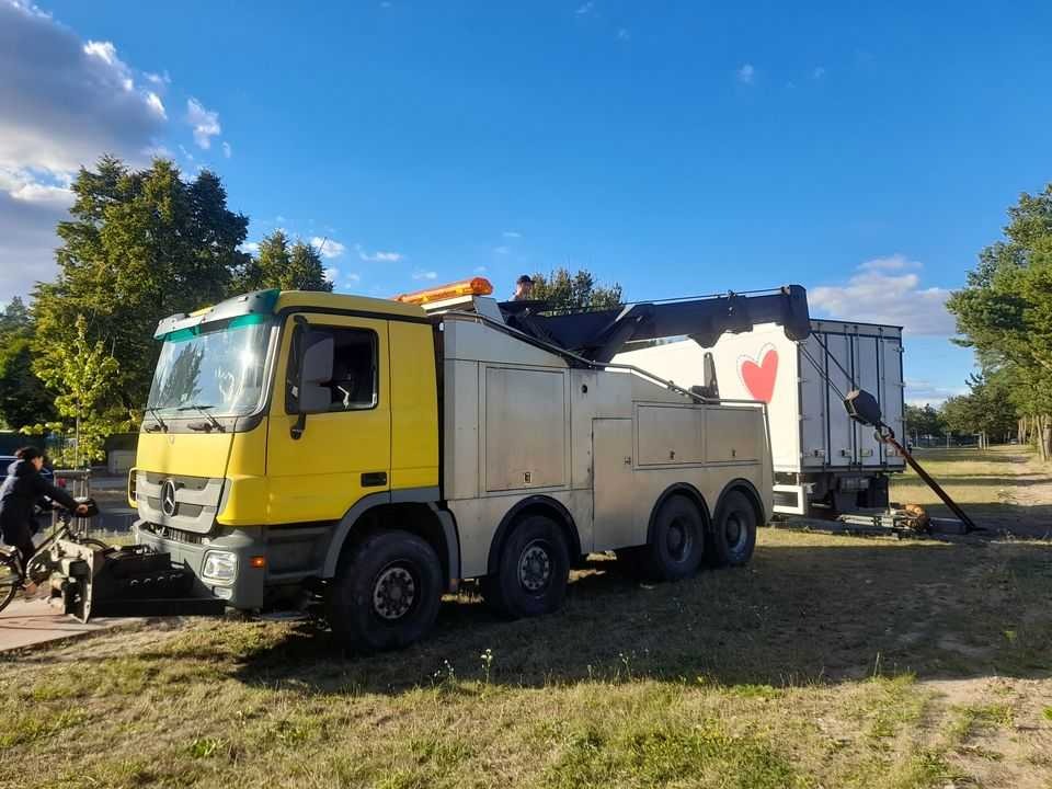 Żółto-srebrny holownik z wysięgnikiem podczepiony do białej naczepy z czerwonym sercem, na tle błękitnego nieba i zieleni drzew.