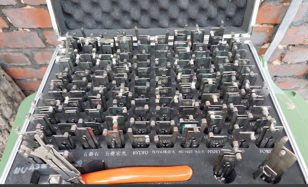 Otwarty futerał z zestawem metalowych kluczy/próbek do zamków samochodowych, ułożonych w rzędach, z pomarańczowym narzędziem widocznym na dole, na tle ceglanej ściany.