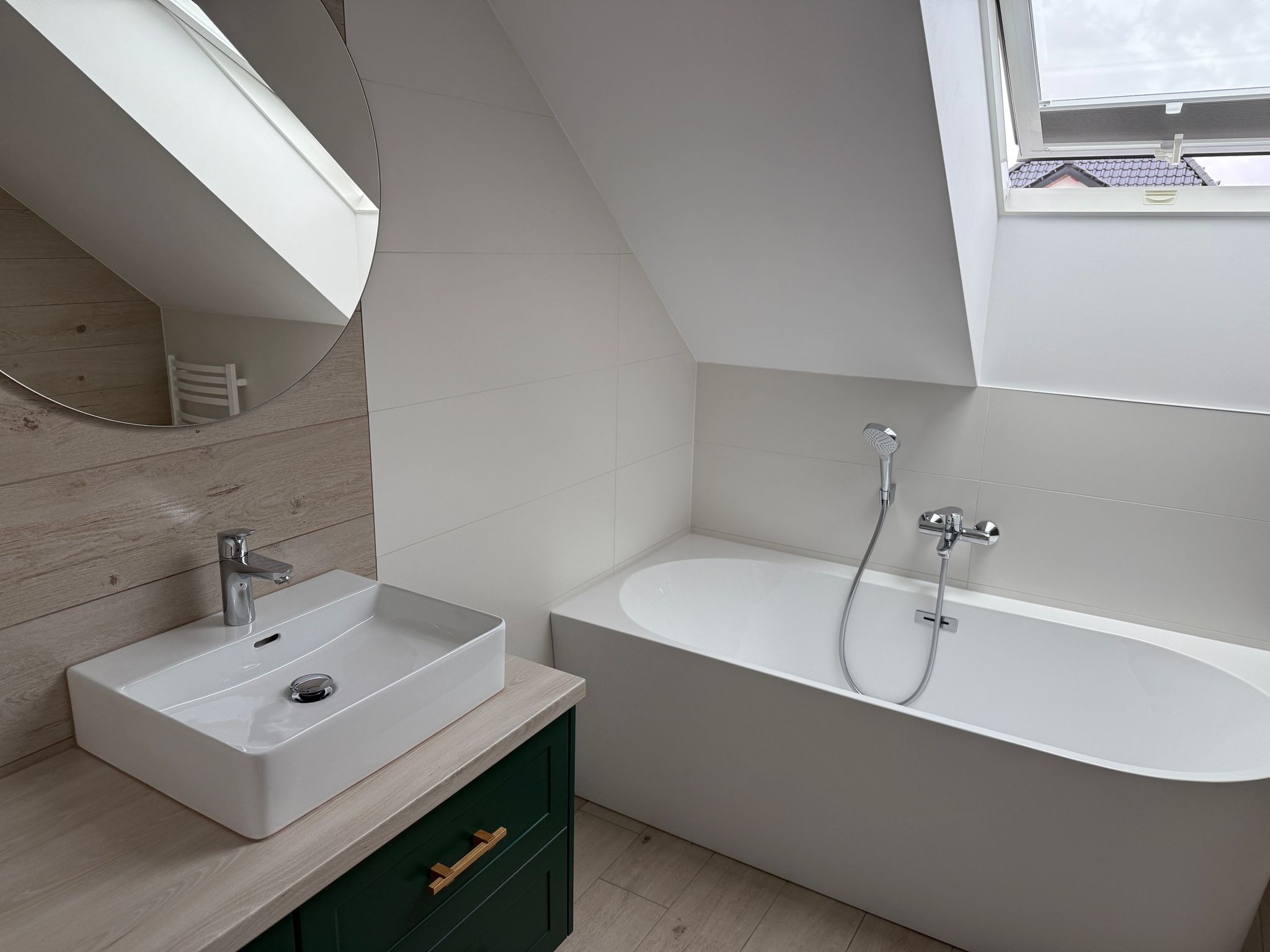 Nowoczesna łazienka na poddaszu z owalną wanną, białą umywalką nablatową na szafce w kolorze butelkowej zieleni i okrągłym lustrem odbijającym grzejnik drabinkowy.