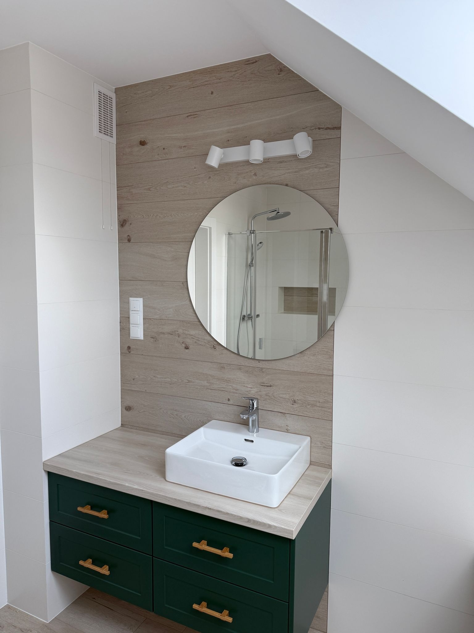 Nowoczesna łazienka na poddaszu z umywalką nablatową, szafką w kolorze butelkowej zieleni z drewnianymi uchwytami, okrągłym lustrem i oświetleniem punktowym.