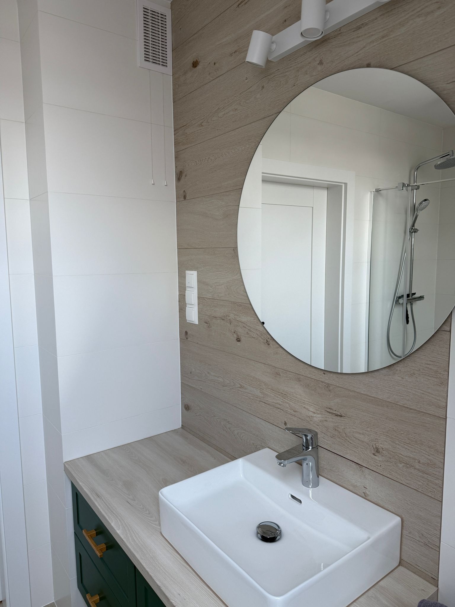 Nowoczesna łazienka z białymi płytkami i drewnopodobną okładziną, okrągłe lustro odbija kabinę prysznicową, pod umywalką ciemnozielona szafka z drewnianym blatem.
