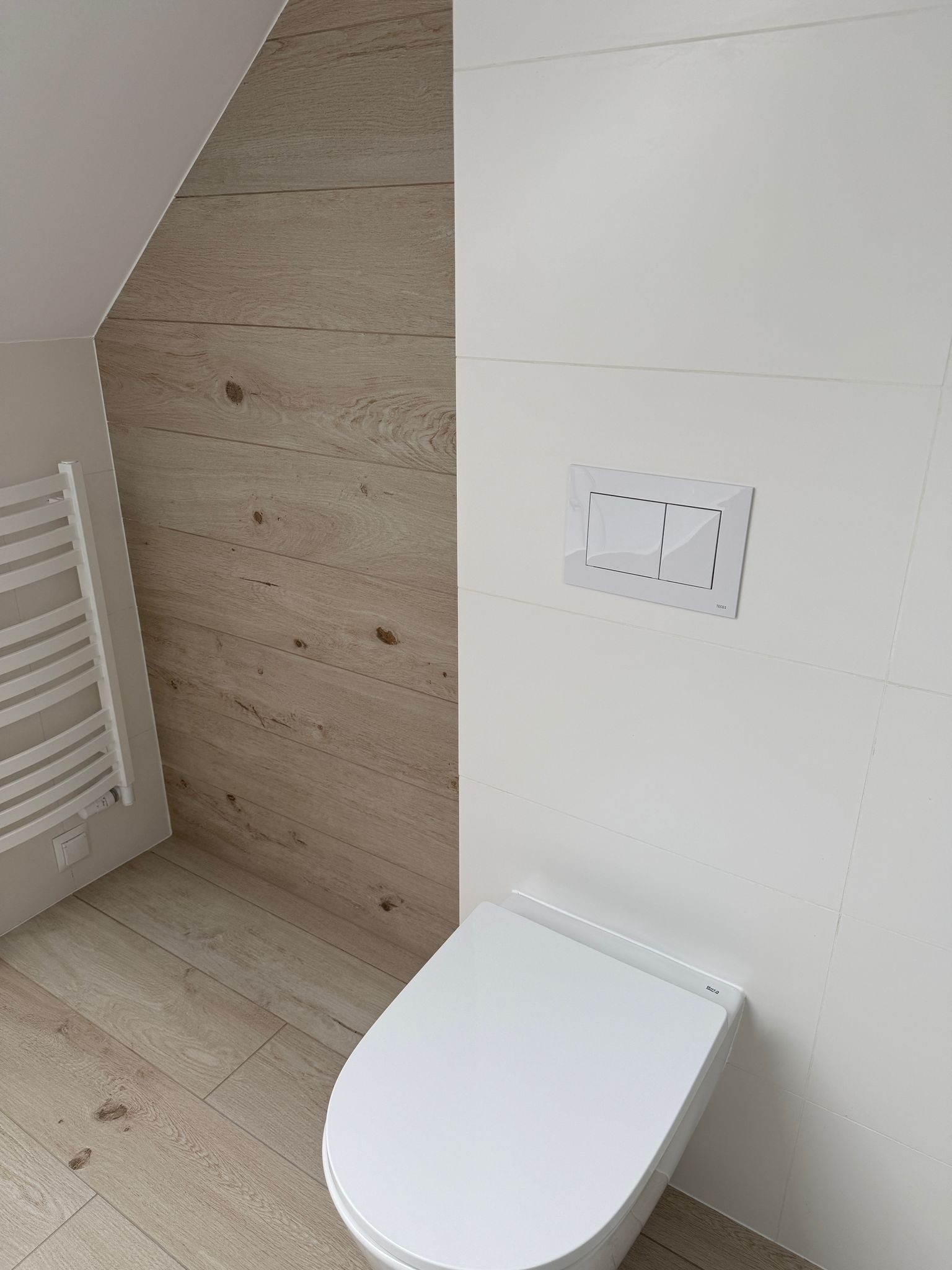 Nowoczesna łazienka z toaletą podwieszaną, białe płytki i drewnopodobne panele ścienne, grzejnik drabinkowy po lewej stronie.