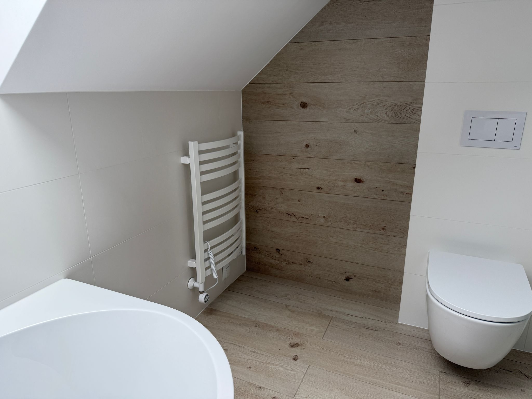 Nowoczesna łazienka na poddaszu z drewnopodobnymi płytkami na podłodze i ścianie, biała armatura i grzejnik drabinkowy.