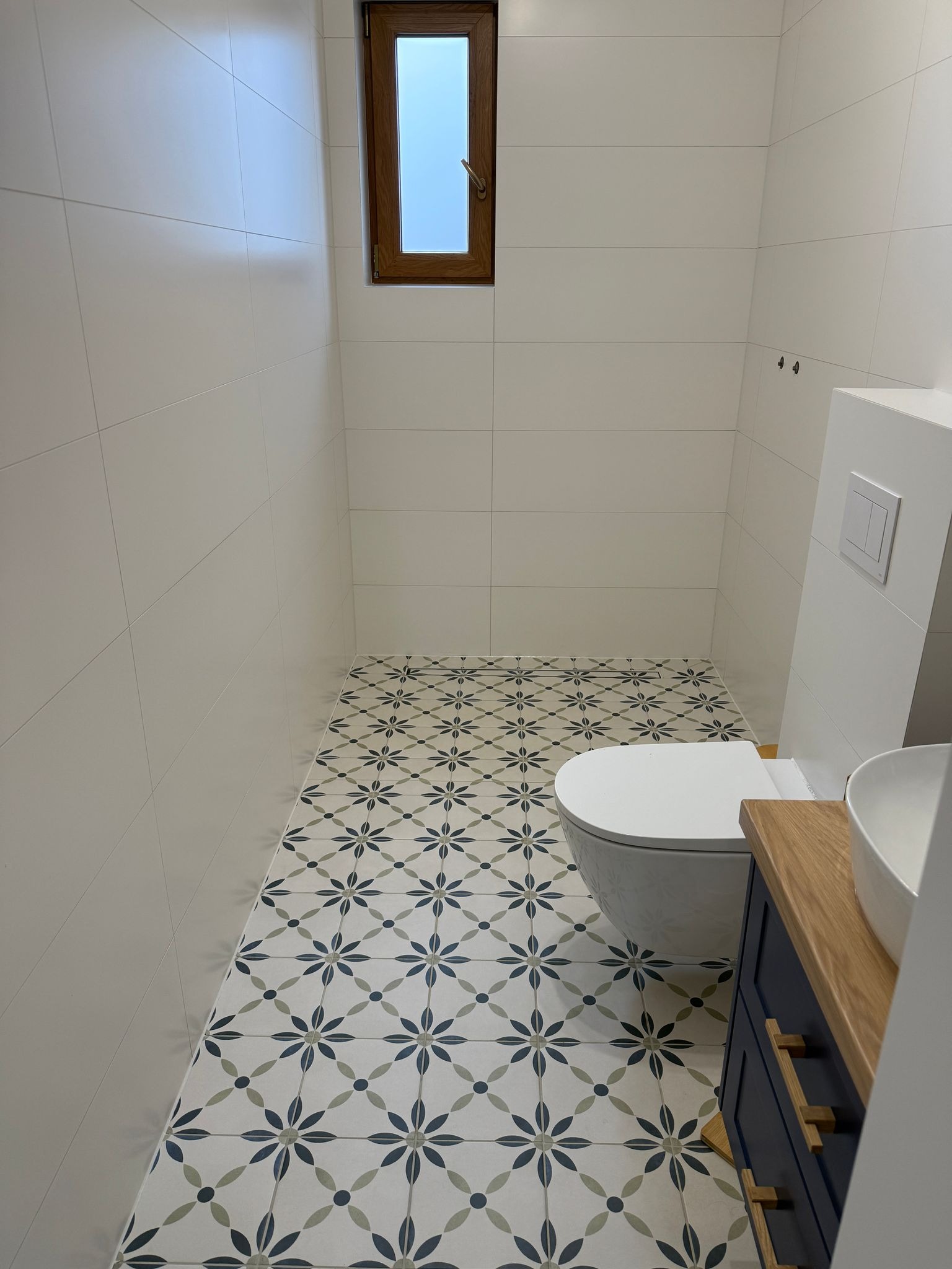 Nowoczesna łazienka z białymi płytkami ściennymi, wzorzystą podłogą w kwiaty, podwieszaną toaletą i szafką pod umywalką w kolorze granatowym z drewnianym blatem.