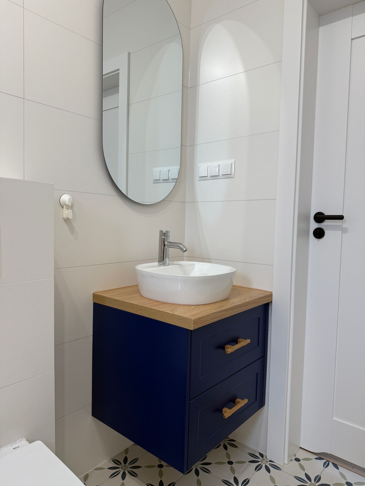 Nowoczesna łazienka z owalnym lustrem, białą umywalką nablatową na szafce w kolorze granatowym z drewnianym blatem, białe płytki ścienne i wzorzysta podłoga.