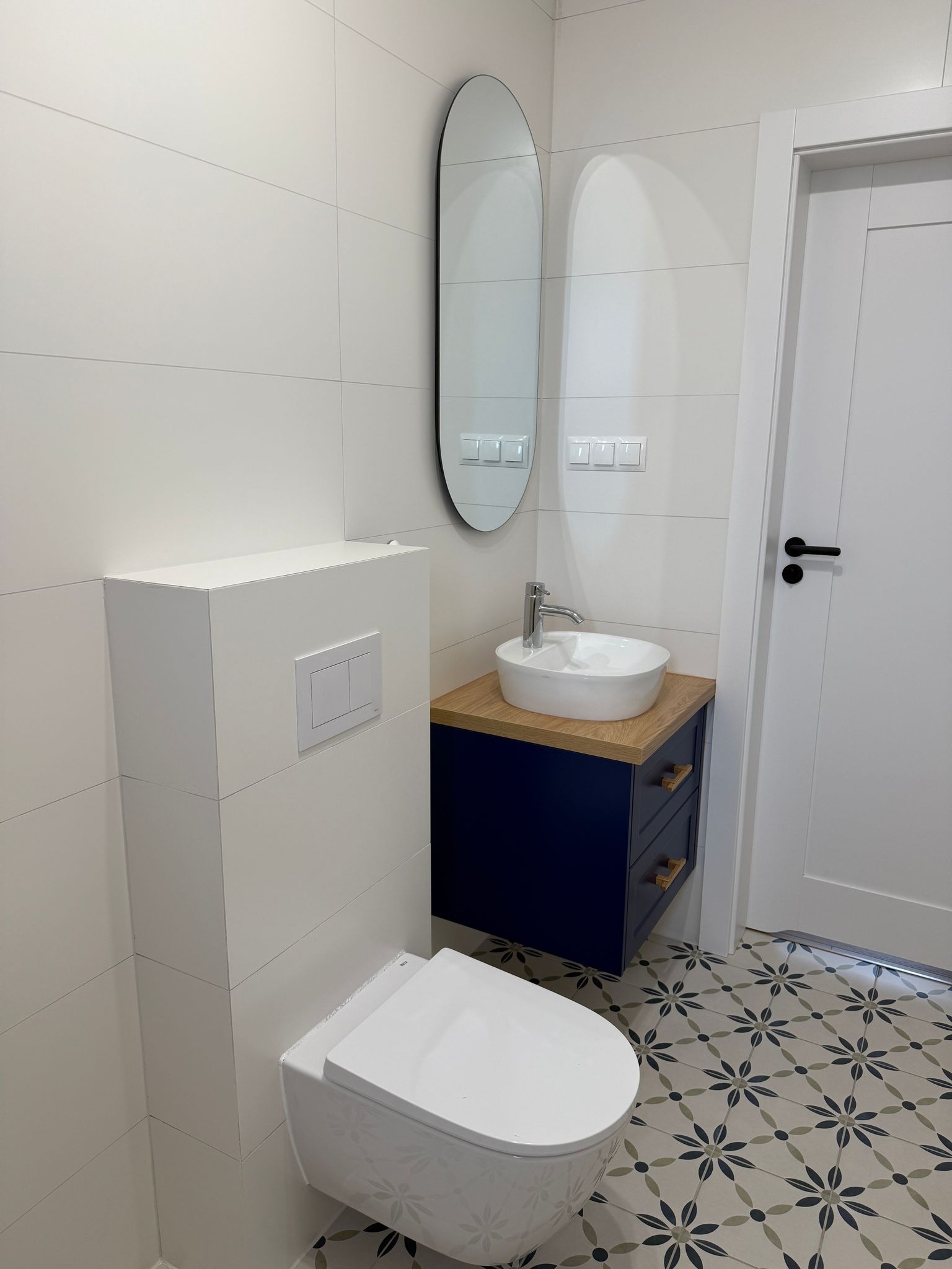 Nowoczesna łazienka z wiszącą toaletą, umywalką nablatową na szafce w kolorze granatowym z drewnianym blatem, owalnym lustrem i wzorzystą podłogą.
