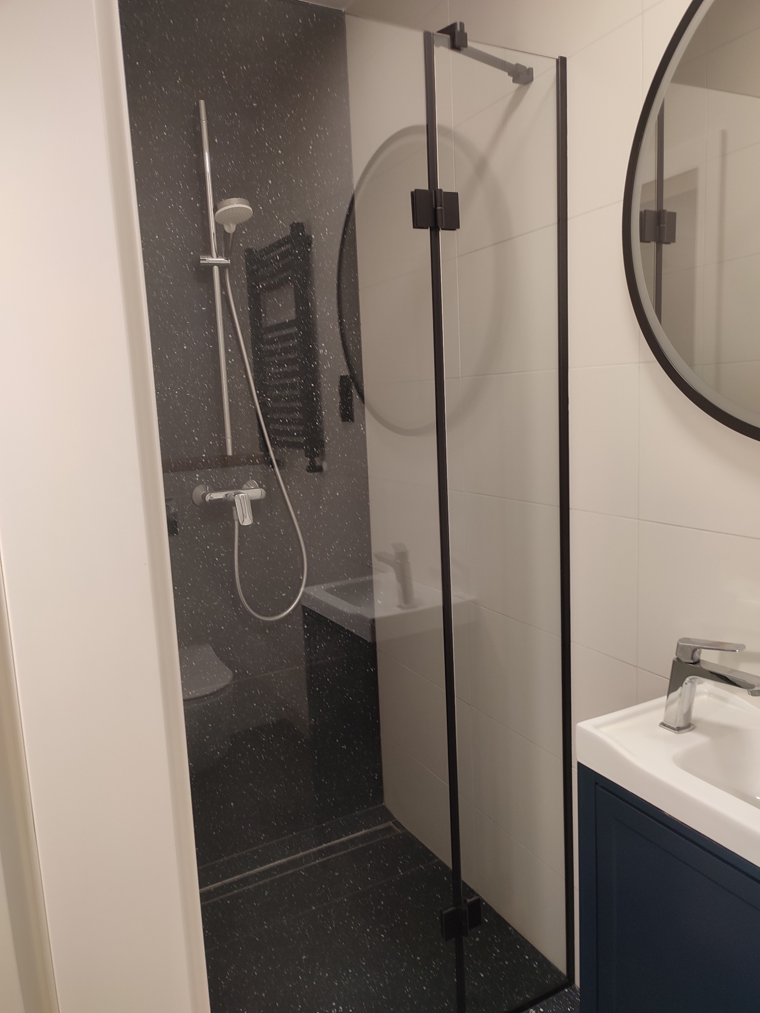 Nowoczesna łazienka z kabiną prysznicową typu walk-in z czarnymi profilami, okrągłym lustrem w czarnej ramie i umywalką na szafce w kolorze granatowym.