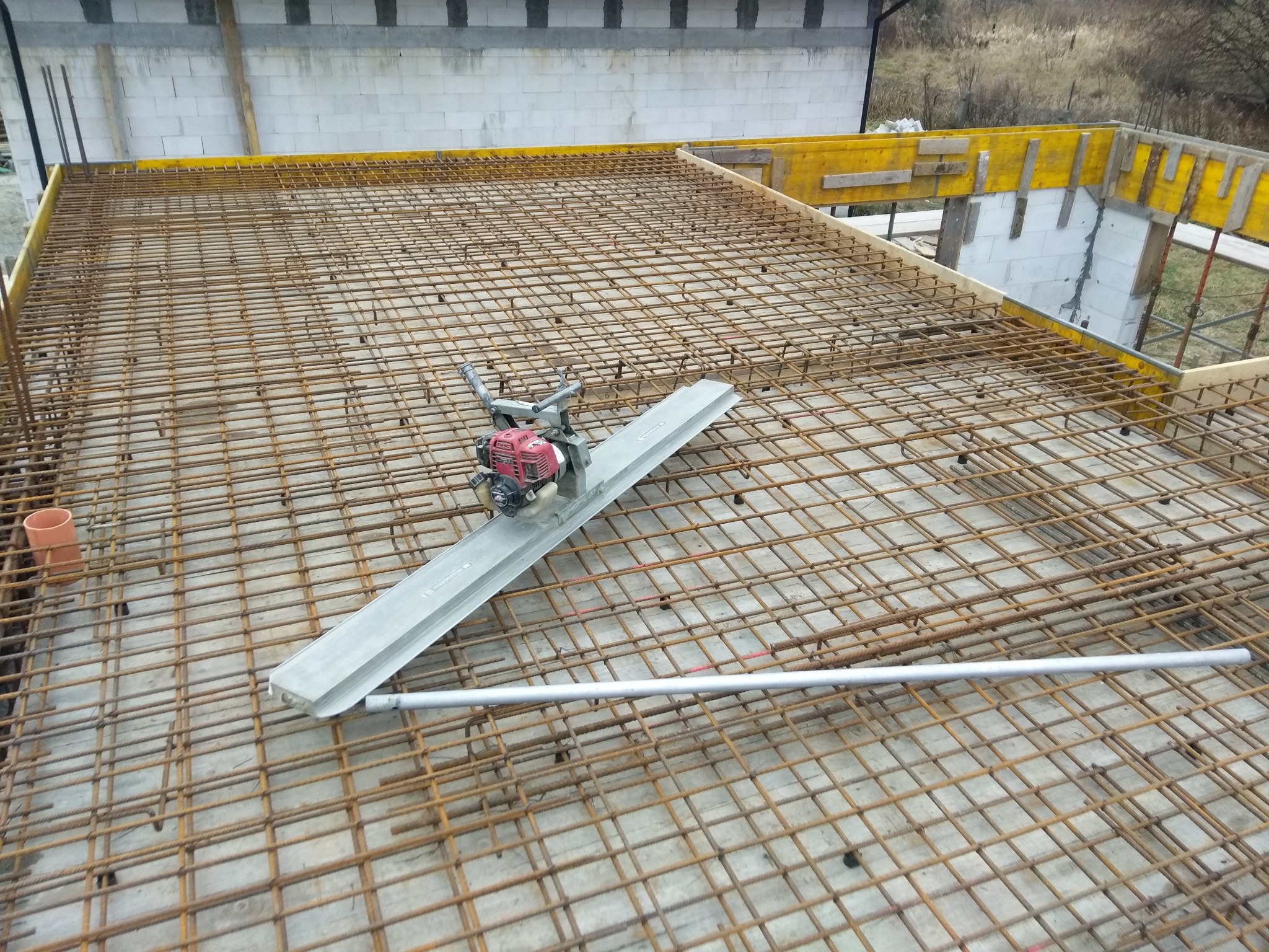 Zbrojenie stropu z siatki stalowej, widoczna listwa wibracyjna do betonu i szalunki. W tle budynek z białego bloczka.