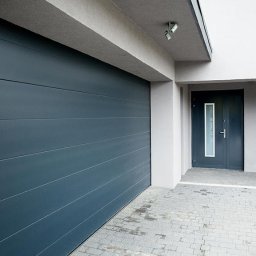 REKO SPÓŁKA Z OGRANICZONĄ ODPOWIEDZIALNOŚCIĄ - Nowoczesna, segmentowa brama garażowa w kolorze antracytowym z poziomymi przetłoczeniami, zamontowana w jasnoszarym budynku z kostki brukowej na podjeździe i małą rośliną ozdobną przy wejściu.