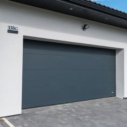 REKO SPÓŁKA Z OGRANICZONĄ ODPOWIEDZIALNOŚCIĄ - Nowoczesna, segmentowa brama garażowa w kolorze grafitowym, zamontowana w jasnoszarym budynku z numerem 135G, widok z zewnątrz na kostkę brukową.