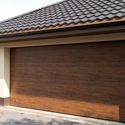 REKO SPÓŁKA Z OGRANICZONĄ ODPOWIEDZIALNOŚCIĄ - Zamknięta, segmentowa brama garażowa w kolorze ciemnego drewna, z widocznym fragmentem dachu z brązowej dachówki ceramicznej i elewacją w odcieniach beżu i brązu.