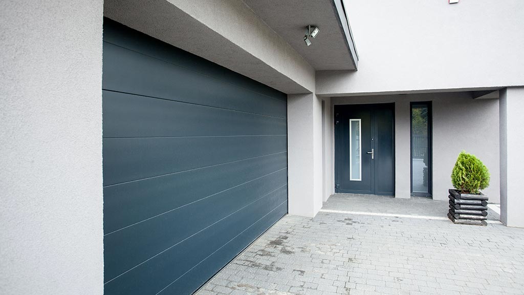 Nowoczesna, segmentowa brama garażowa w kolorze antracytowym z poziomymi przetłoczeniami, zamontowana w jasnoszarym budynku z kostki brukowej na podjeździe i małą rośliną ozdobną przy wejściu.