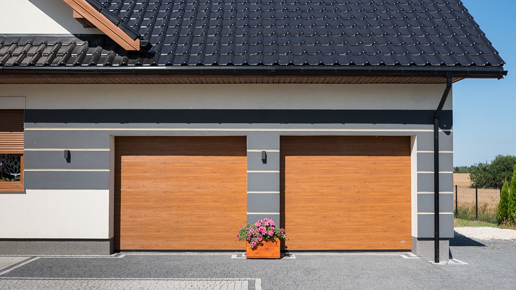 Elewacja domu z dwoma drewnopodobnymi bramami garażowymi, czarnym dachem z dachówki i skrzynką z różowymi kwiatami stojącą przed bramami.
