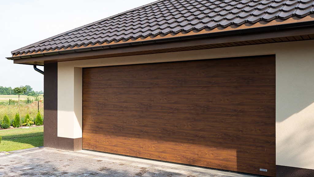 Zamknięta, segmentowa brama garażowa w kolorze ciemnego drewna, z widocznym fragmentem dachu z brązowej dachówki ceramicznej i elewacją w odcieniach beżu i brązu.