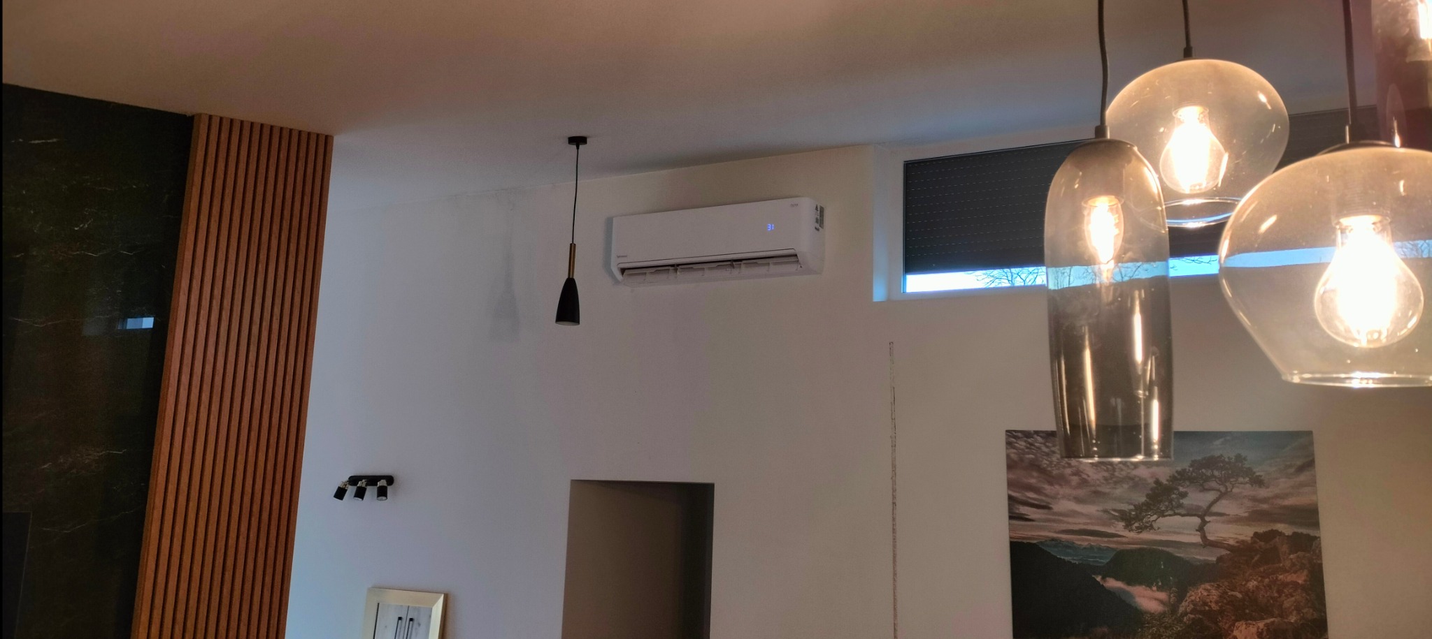Biała jednostka wewnętrzna klimatyzacji zamontowana na ścianie w nowoczesnym salonie z drewnianymi panelami, oświetleniem punktowym i dekoracyjnymi lampami wiszącymi.
