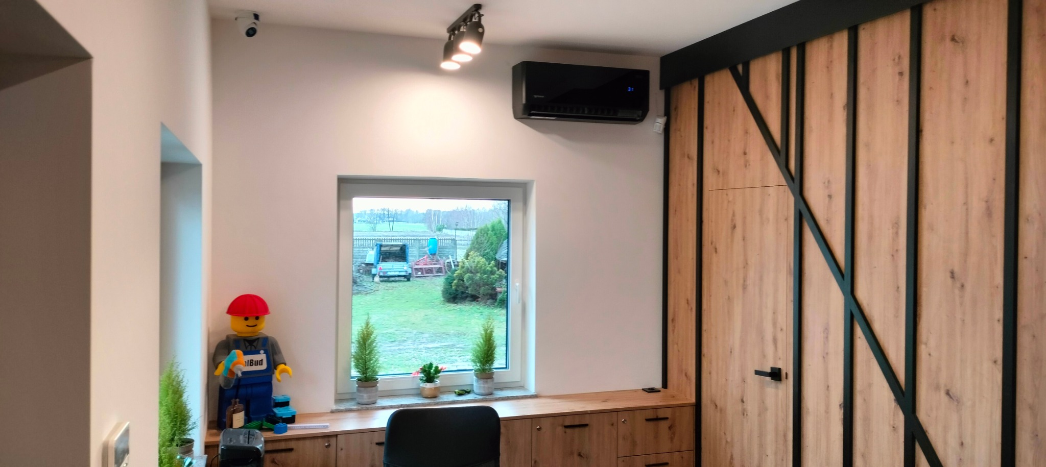 Czarna jednostka klimatyzacyjna zamontowana na ścianie w nowoczesnym wnętrzu, z widokiem na okno i dekoracyjną szafą z drewnianymi frontami oraz figurką Lego w stroju budowlanym.