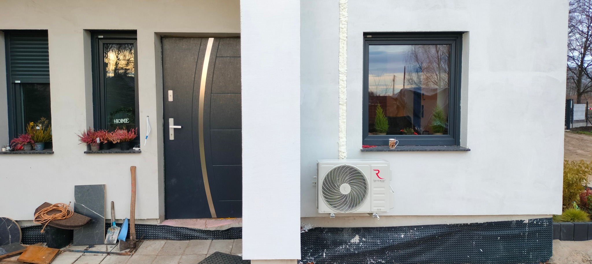 Biała zewnętrzna jednostka klimatyzacji Rotenso zamontowana na ścianie domu obok okna z roślinami i kubkiem, widok na drzwi wejściowe, narzędzia i materiały budowlane.