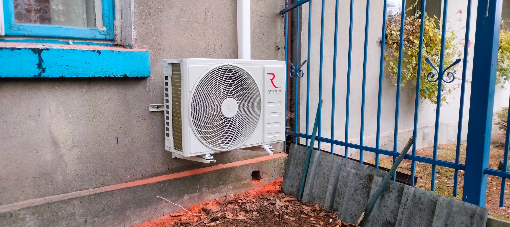 Biała jednostka zewnętrzna klimatyzatora marki Rotenso zamontowana na szarej ścianie budynku, widoczna niebieska rama okienna i niebieskie metalowe ogrodzenie z ozdobnymi elementami.