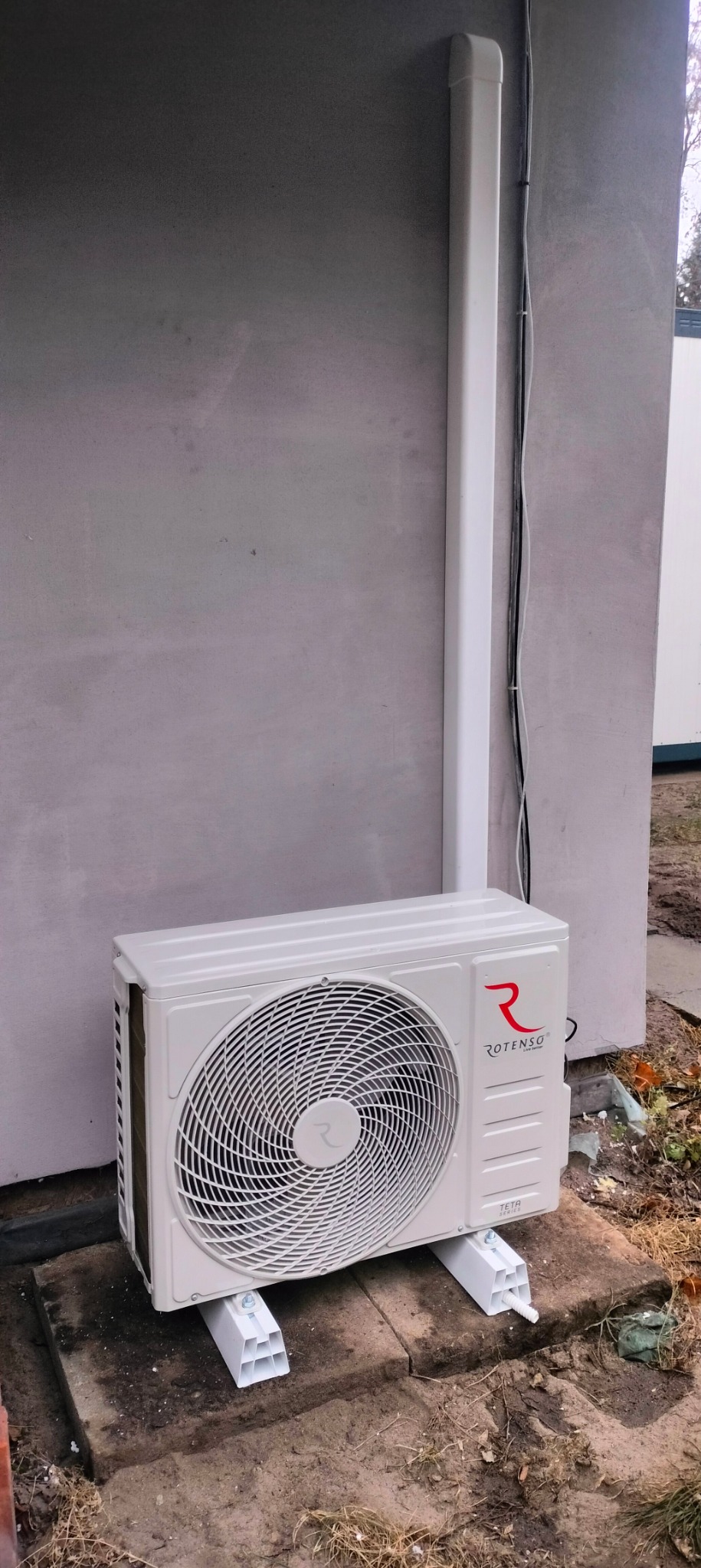 Zewnętrzna jednostka klimatyzacji Rotenso TETA Series zamontowana na białych wspornikach z odprowadzeniem skroplin, przy szarej ścianie budynku.