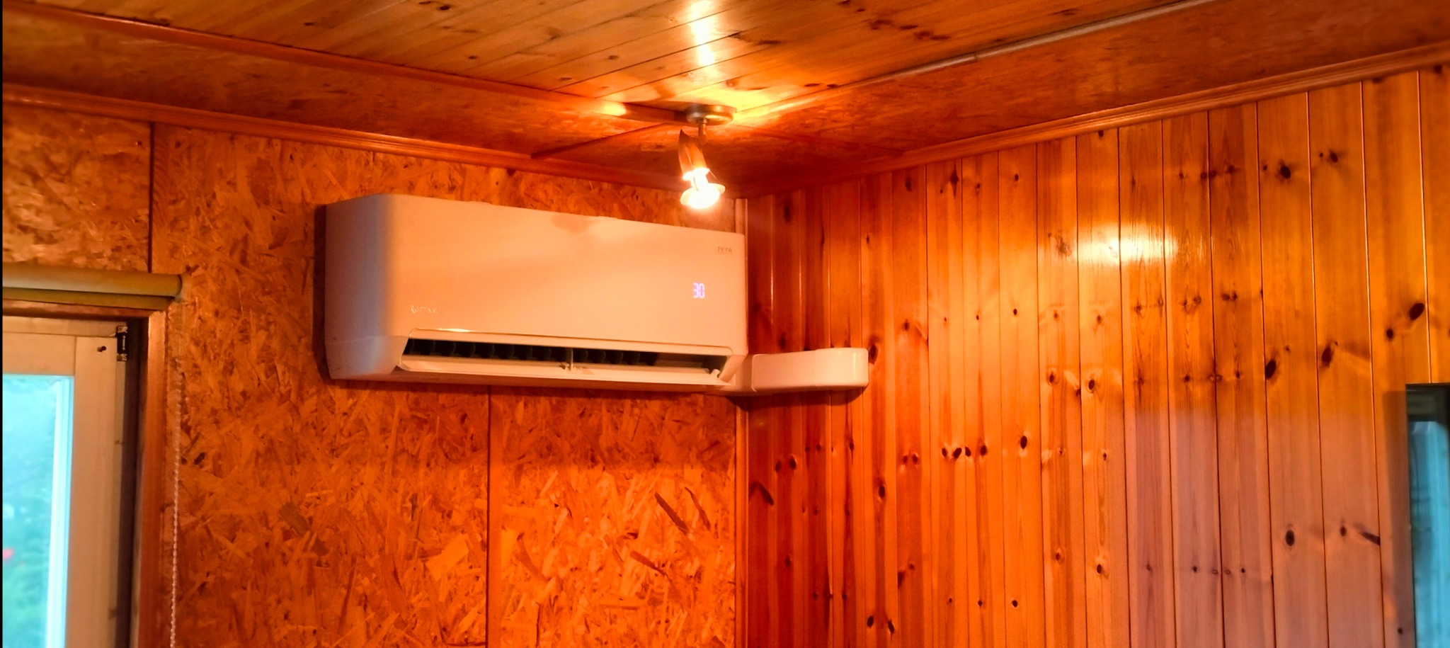 Klimatyzator ścienny w drewnianym wnętrzu, ustawiony na 30 stopni, oświetlony ciepłym światłem żarówki.
