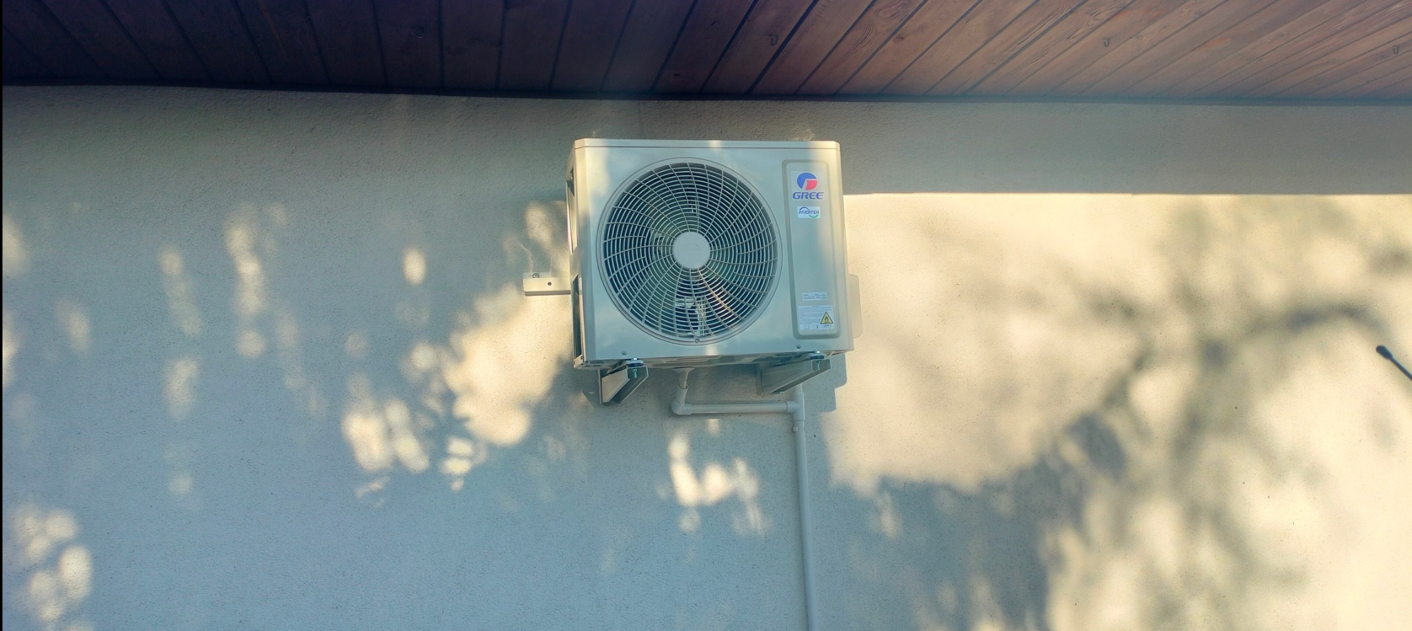 Zewnętrzna jednostka klimatyzacji marki Gree zamontowana na jasnej ścianie budynku, oświetlona popołudniowym słońcem, rzucająca cienie liści na elewację.