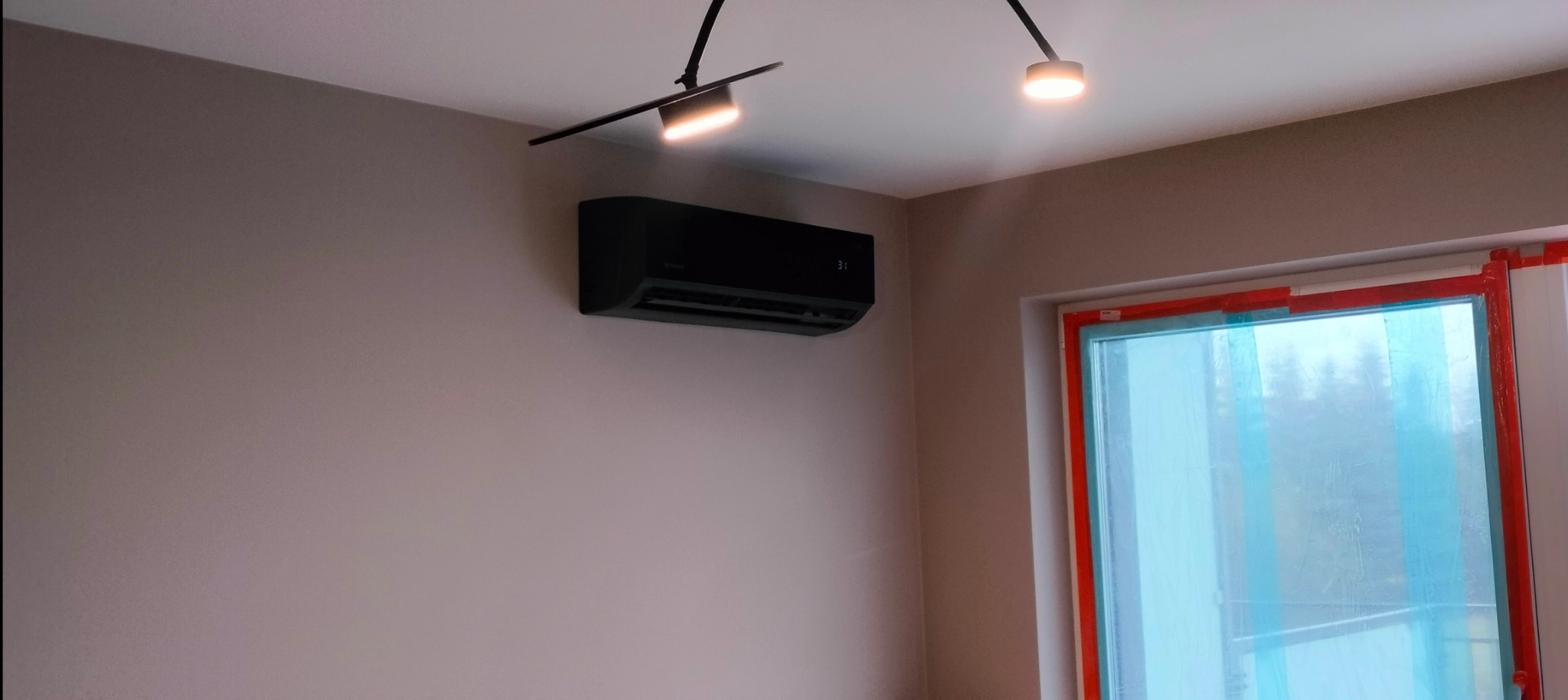 Czarna jednostka klimatyzacji ściennej, zamontowana w pokoju z szarą ścianą i oknem zabezpieczonym folią ochronną, oświetlona dwoma reflektorami sufitowymi.